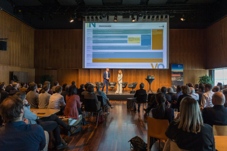 Die INNOVATE VORARLBERG mit Publikum, den zwei Sprechenden Kerstin Polzer und Jimmy Heinzl auf der Bühne und einer großen Programmübersicht im Hintergrund.