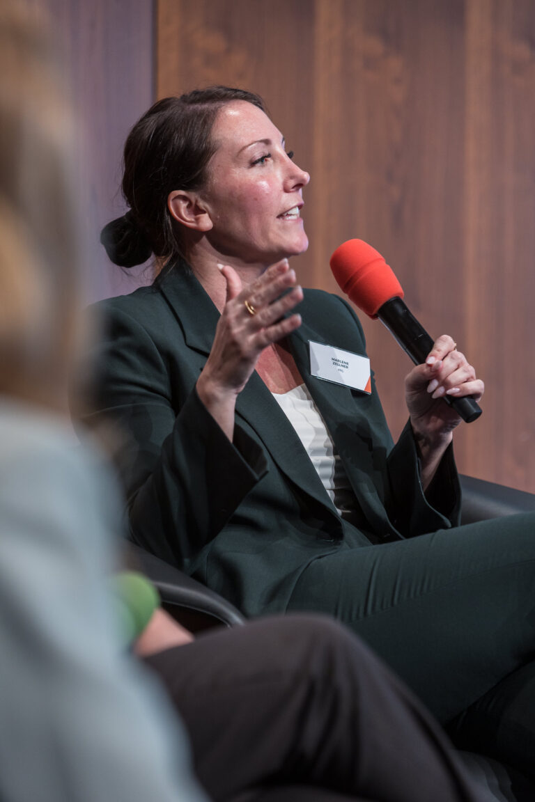 Eine spricht auf eine Bühne sitzend mit einem Mikrofon