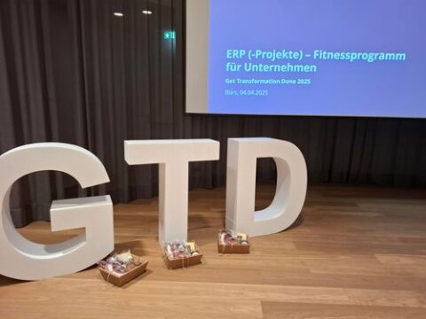 Große weiße Buchstaben „G T D“ stehen vor einer Leinwand, auf der eine Präsentation zum Thema ERP-Projekte gezeigt wird.