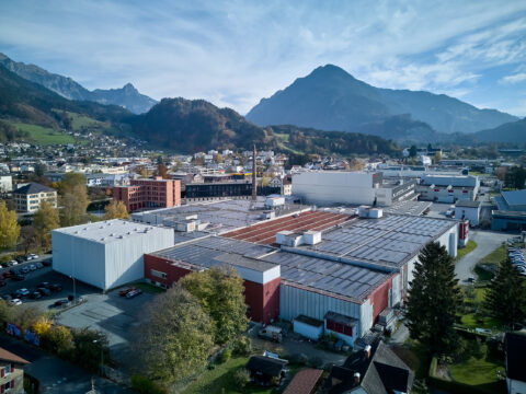 Luftaufnahme des Getzner Textil Firmengeländes in Bludenz mit mehreren Fabrikgebäuden und einem hohen Schornstein. Auf den Dächern sind großflächige Photovoltaikanlagen installiert, im Hintergrund sind Berge zu sehen.
